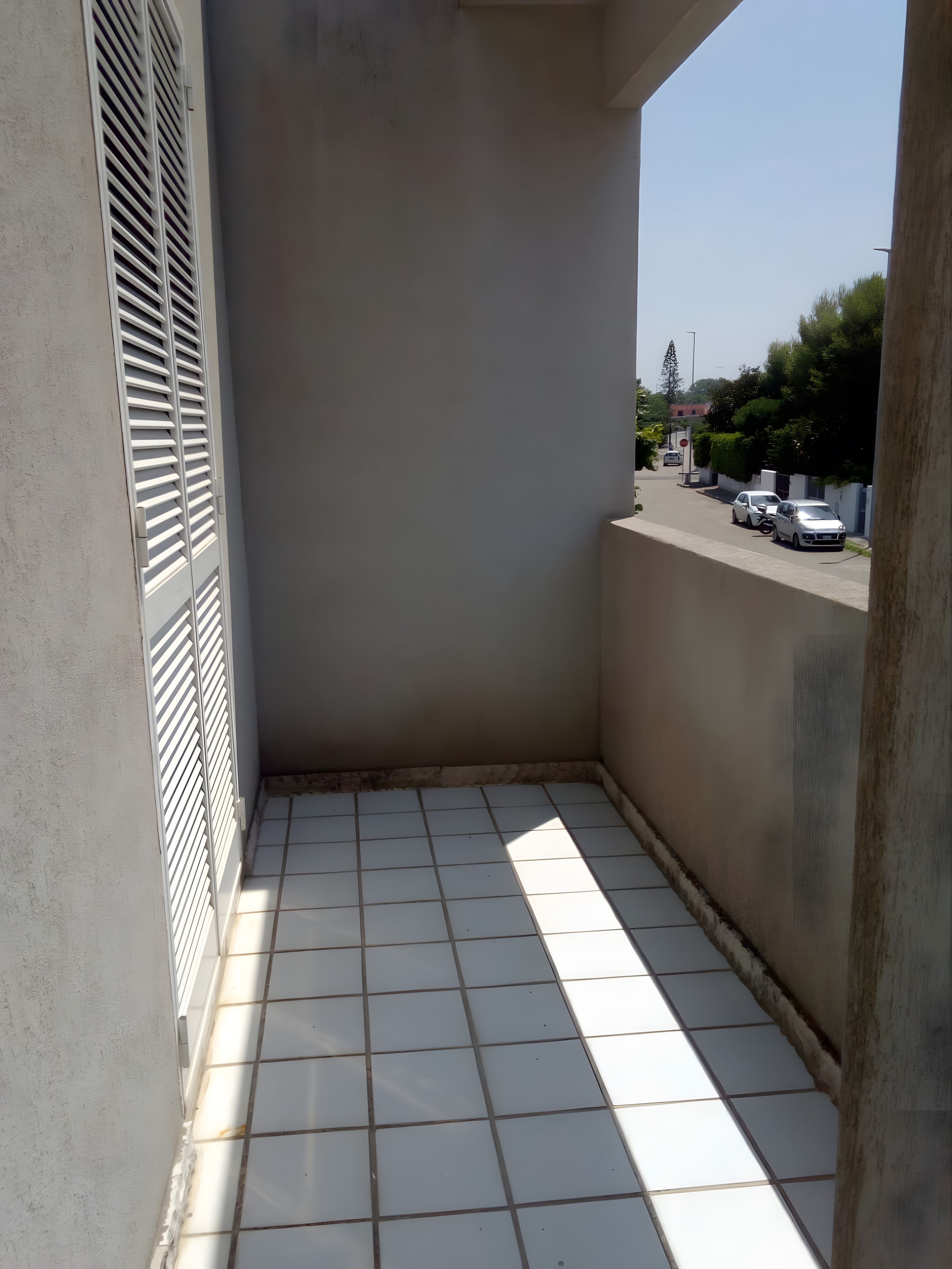 A balcony with a tiled floor, a metal grill on the left, and a view of parked cars and greenery in the background.