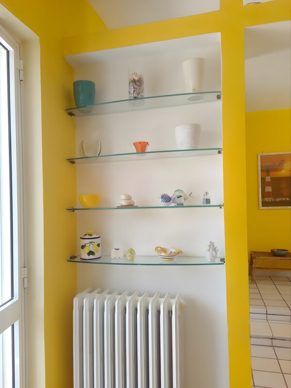 Habitació amb un radiador blanc, prestatges de vidre amb diverses eines de cuina i una paret groga.