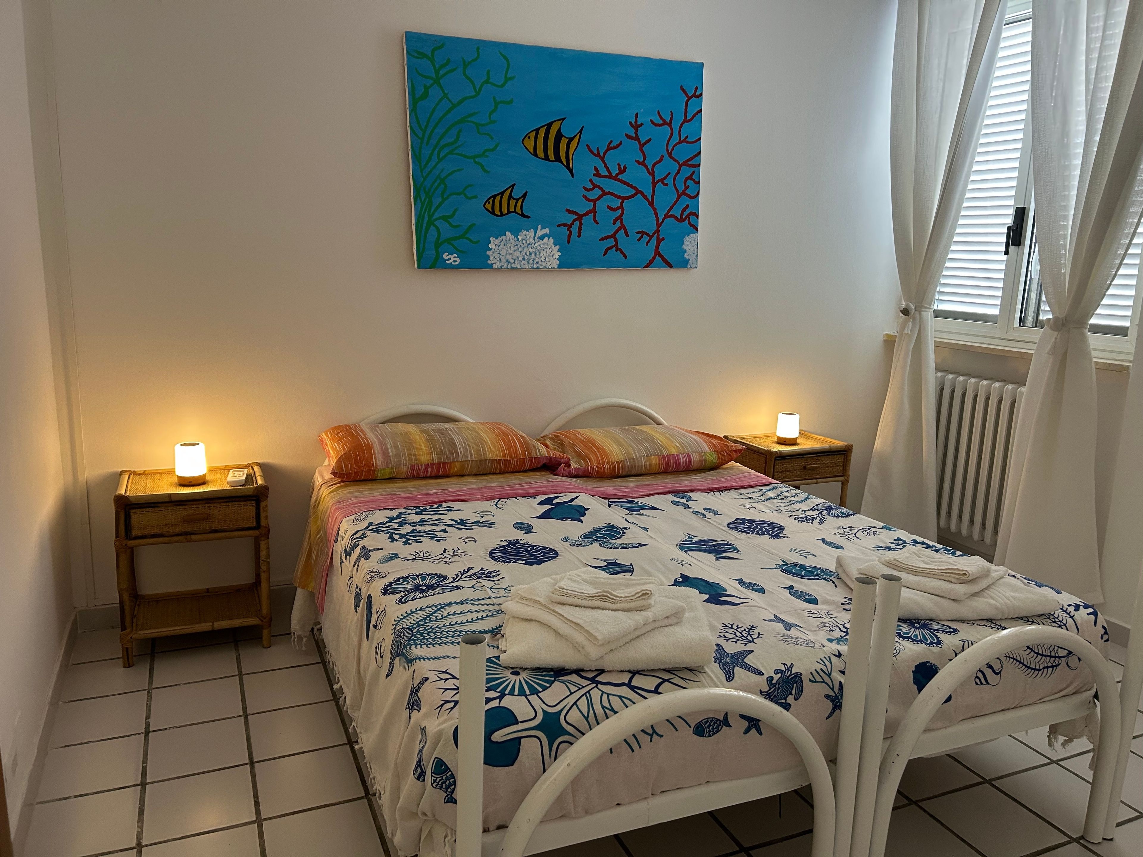 A bedroom with a bed, two bedside lamps, a painting, and a window with curtains.