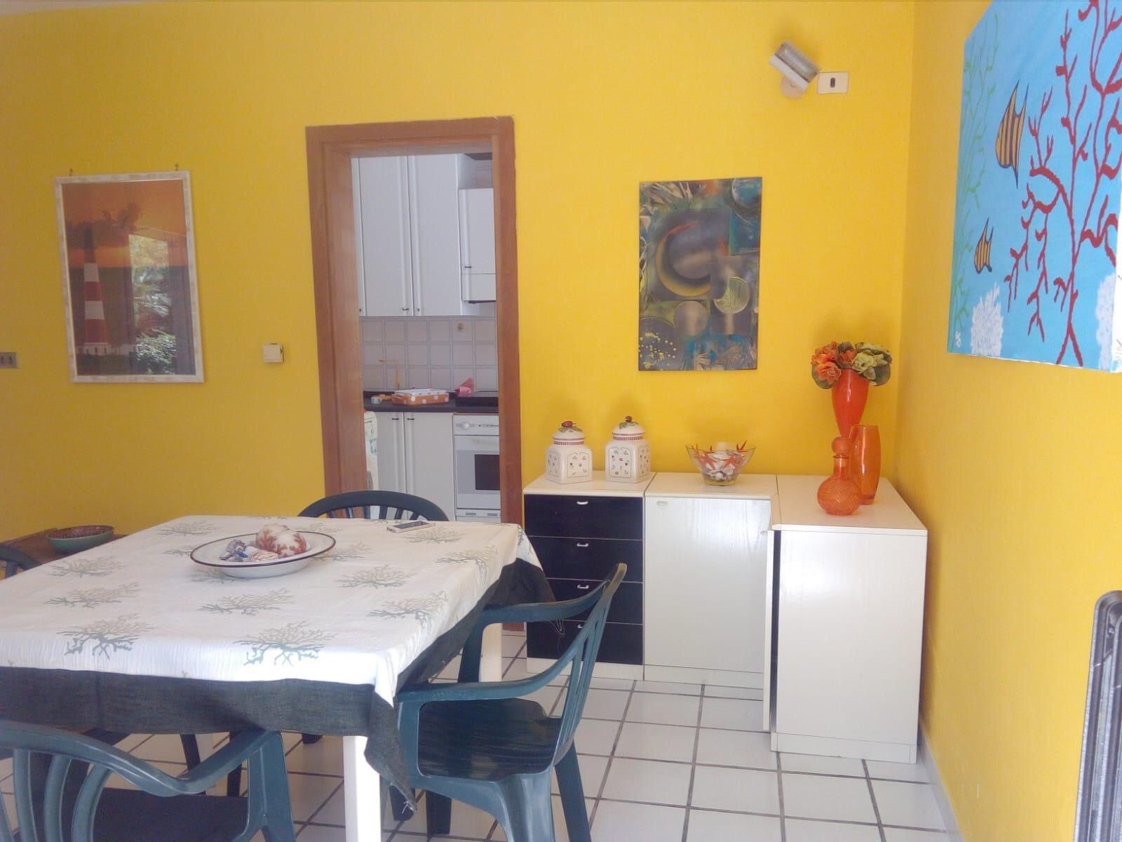 Cozinha compacta com armários brancos, mesa de jantar com cadeiras e uma geladeira pequena.