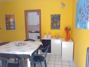 Cozinha compacta com armários brancos, mesa de jantar com cadeiras e uma geladeira pequena.
