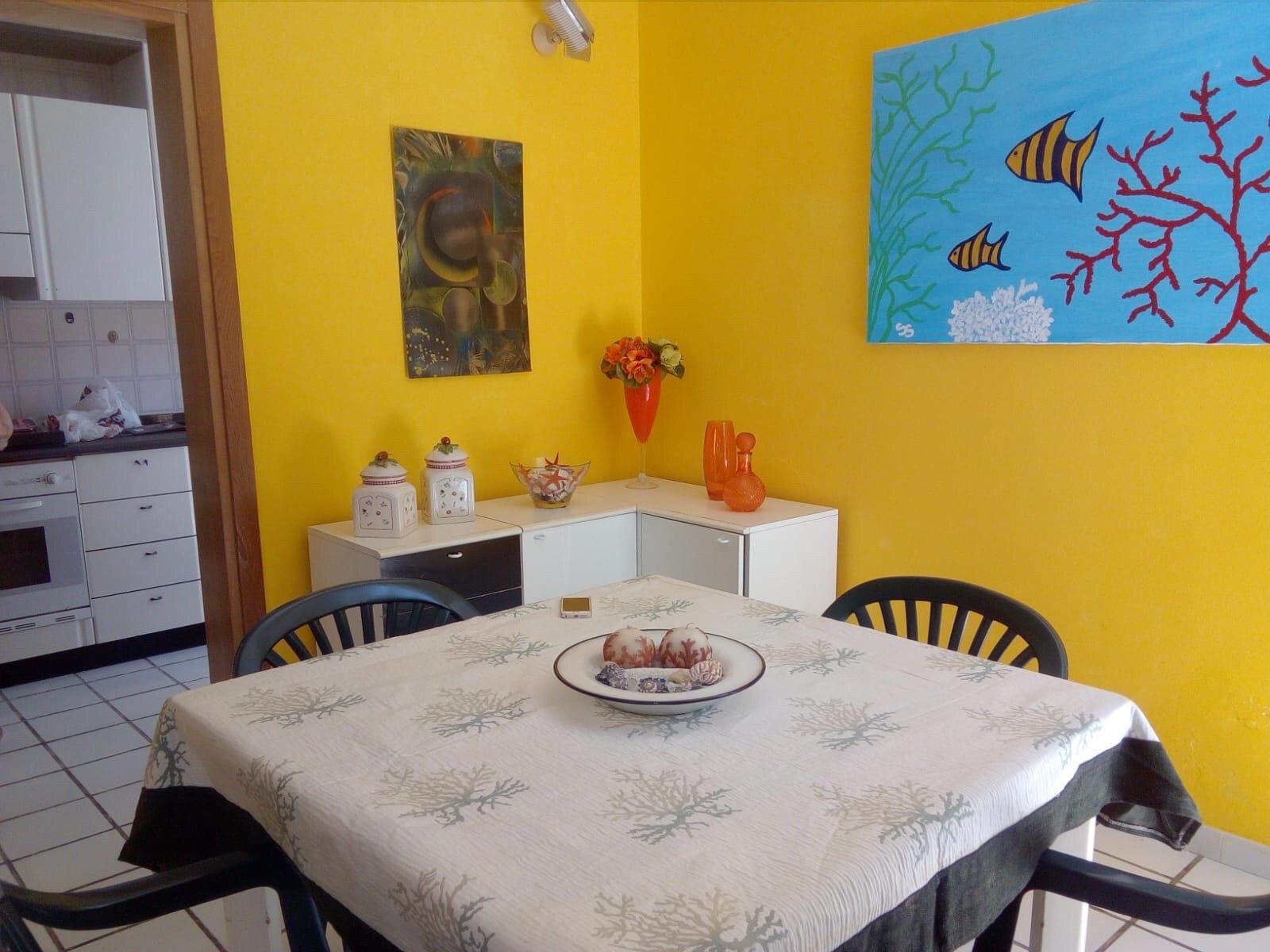 A dining area with a table set for two, a painting above the table, and a kitchen in the background.