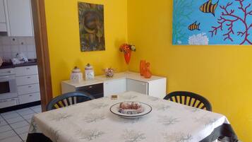 A dining area with a table set for two, a painting above the table, and a kitchen in the background.