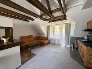 A loft-style living space with a sloped ceiling, a sofa, a coffee table, a kitchen area with wooden cabinets, and a window with curtains.