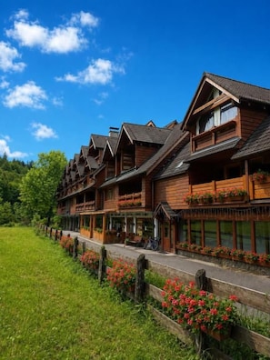 A row of wooden buildings with balconies, a paved walkway, and a grassy area with flowers. - Penzión SIMI (Vysoké Tatry)