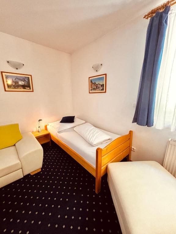 A small, single-bed room with a wooden bed, a white sofa, a small table, and two wall-mounted lamps.