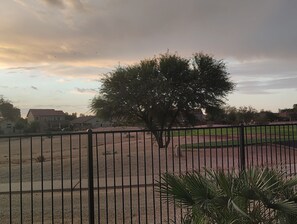 A fenced sports field with a large tree in the foreground. - Enjoy breathtaking sunrises and sunsets on Johnson Ranch golf course and unforgettable stargazing from the peaceful backyard. (San Tan Valley)