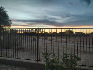 A fenced outdoor area with a gravel surface, surrounded by vegetation and houses in the background. - Enjoy breathtaking sunrises and sunsets on Johnson Ranch golf course and unforgettable stargazing from the peaceful backyard. (San Tan Valley)