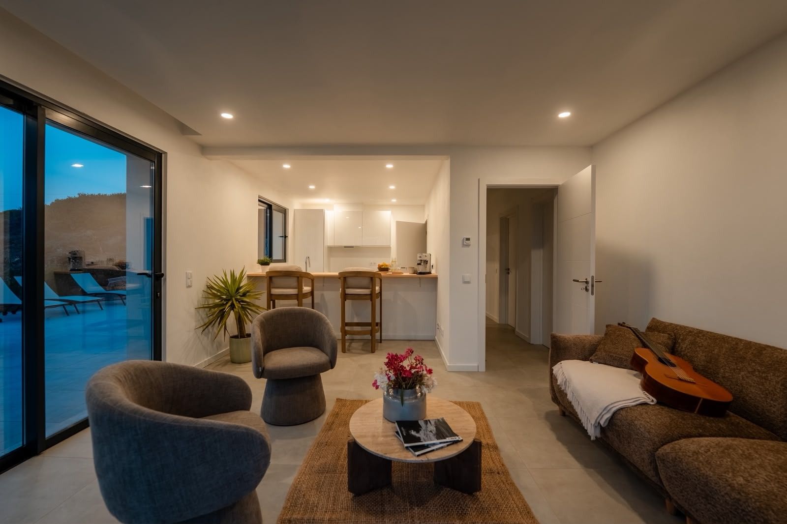 Ein modernes Wohnzimmer mit einer Couch, Sesseln, einem Couchtisch und einem Essbereich mit Stühlen, im Hintergrund eine Küche.