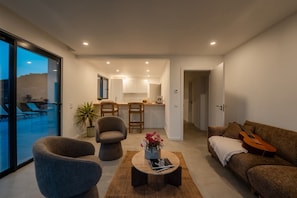 Ein modernes Wohnzimmer mit einer Couch, Sesseln, einem Couchtisch und einem Essbereich mit Stühlen, im Hintergrund eine Küche.