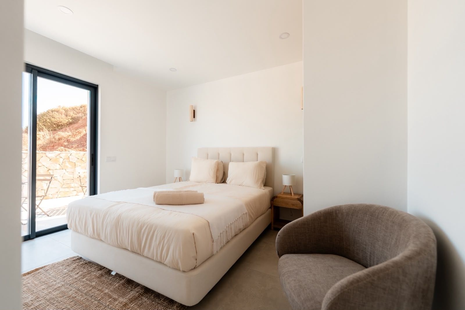 A bedroom with a large bed, a chair, a nightstand, and a view of the outdoors.
