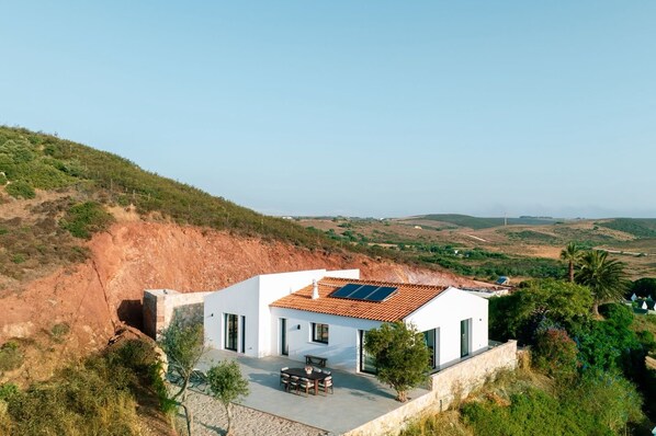 A modern house with a solar panel roof, a patio with a table and chairs, and a view of the surrounding landscape.