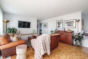 Un salon comprenant un canapé, une table basse et un téléviseur fixé au mur. On y trouve également un coin repas avec une table et des chaises en bois.