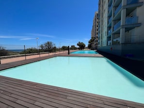 Ein Schwimmbad mit Holzterrasse, umgeben von einem Zaun und einem Gebäude mit Balkonen.