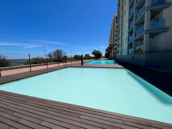 A swimming pool with a wooden deck, surrounded by a fence and a building with balconies.