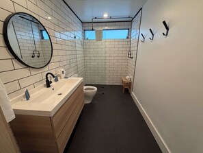Een moderne badkamer met een grote spiegel, witte betegelde wanden, een houten wastafelmeubel, een toilet en een klein houten krukje.