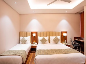 A hotel room with two beds, a desk, a chair, and a ceiling fan. - Deluxe Twin Room Hotel (Agra)