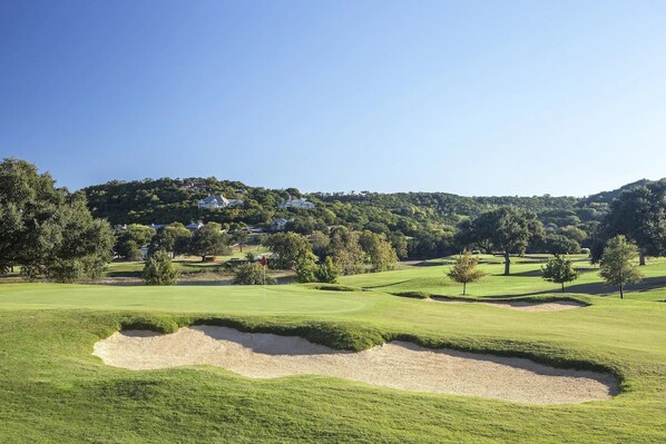 Ein Golfplatz mit Bunkern, Bäumen und Häusern im Hintergrund.