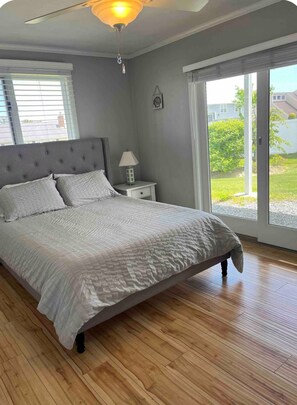 A bedroom with a bed, a nightstand, a lamp, a ceiling fan, and a view of the outdoors. - 🐋 Finback Beach House 🐋 Walk to Scarborough Beach, AC, Firepit, Bright & Open (Narragansett)