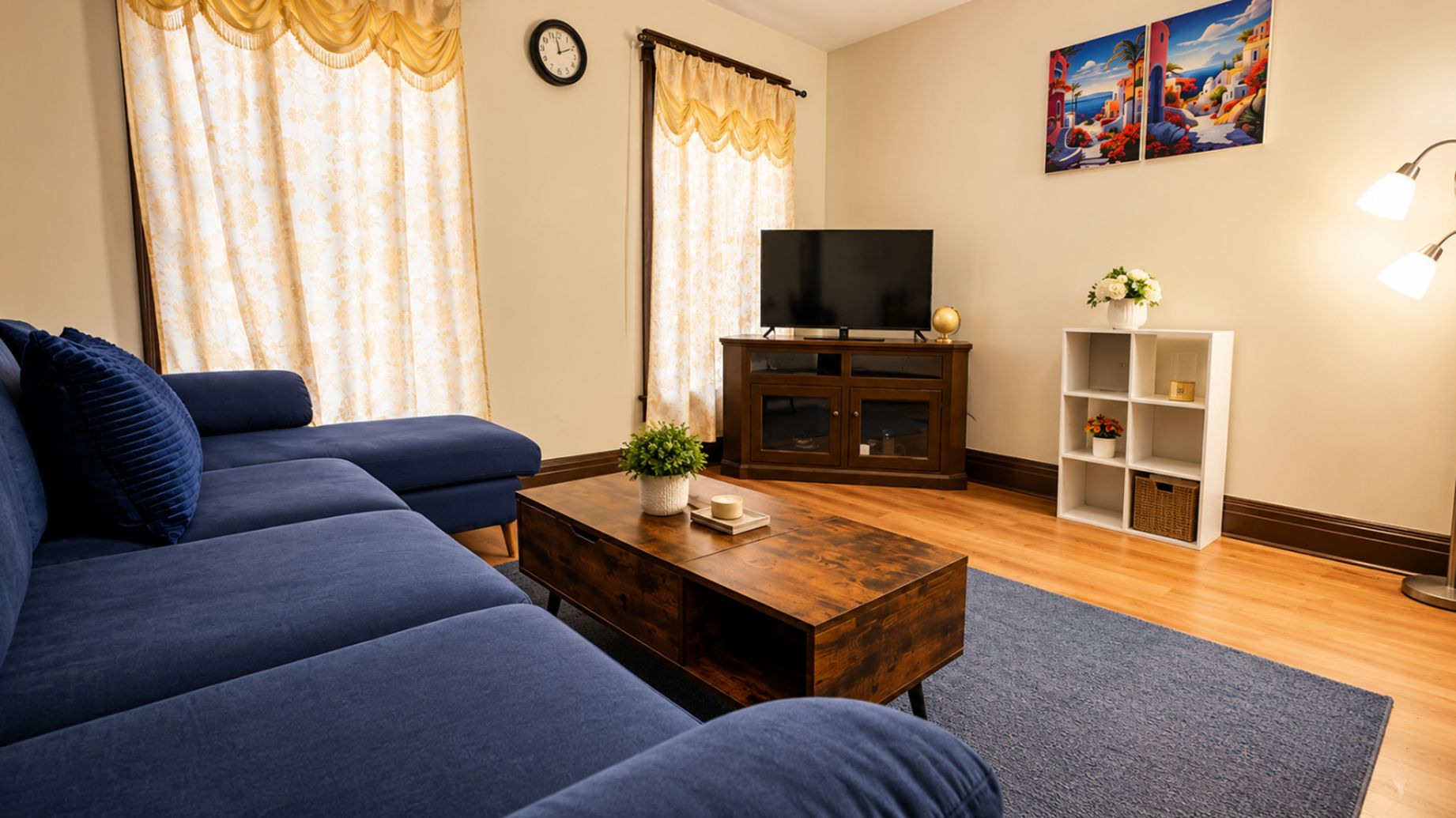 A living room with a blue sofa, a wooden coffee table, a TV on a cabinet, a clock on the wall, and a painting above the TV.