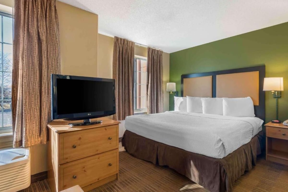 A hotel room with a bed, a television on a wooden dresser, a nightstand with a lamp, and a view of the outdoors through the window.