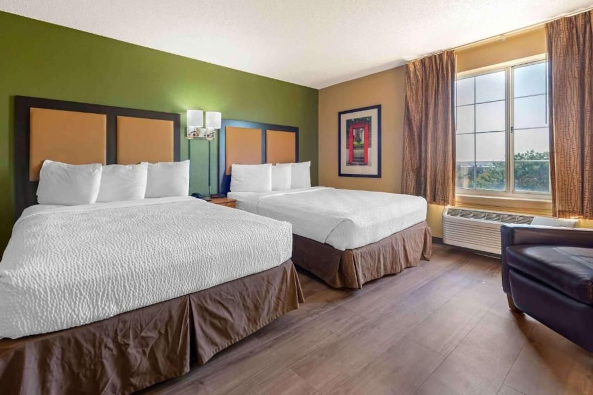 A hotel room with two beds, a window with curtains, a chair, and a wall-mounted air conditioner.