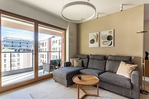 A modern living room with a sectional sofa, a round wooden coffee table, and a view of a cityscape through large sliding glass doors. - Towarowa Square V P&O Apartments (Warszawa)
