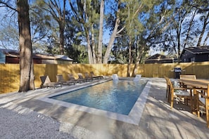 Piscina retangular com deck de madeira ao redor, equipada com espreguiçadeiras e uma mesa de jantar.