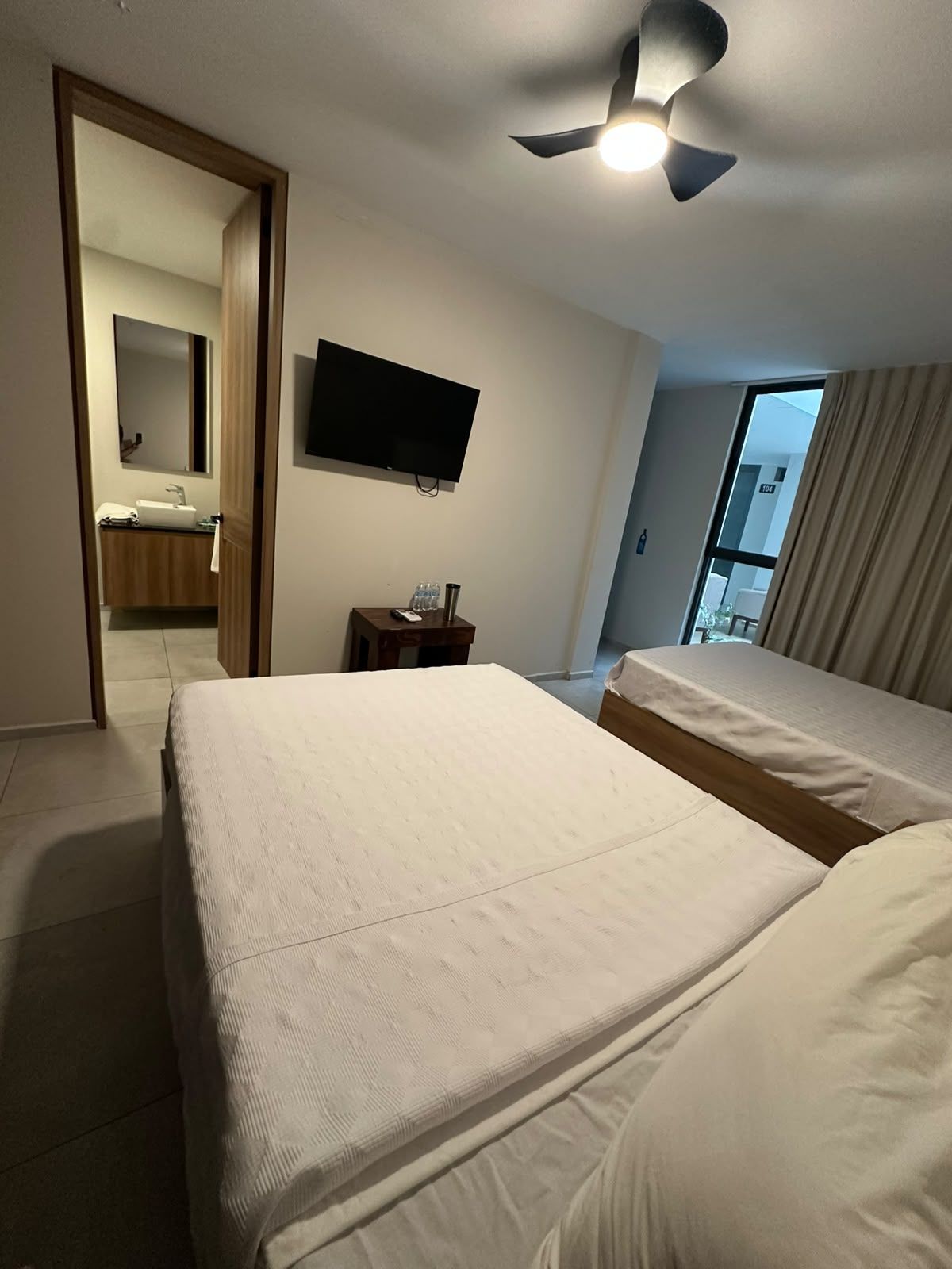 Camera d'albergo con due letti, una televisione, una ventola a soffitto, un bagno parzialmente visibile attraverso una porta aperta e una finestra con tende.
