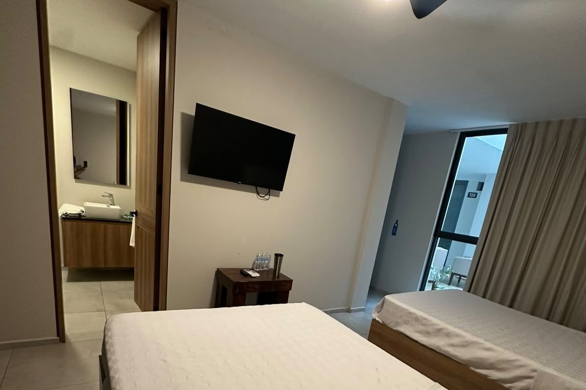 A hotel room with two beds, a television, a ceiling fan, a bathroom visible through an open door, and a window with curtains.