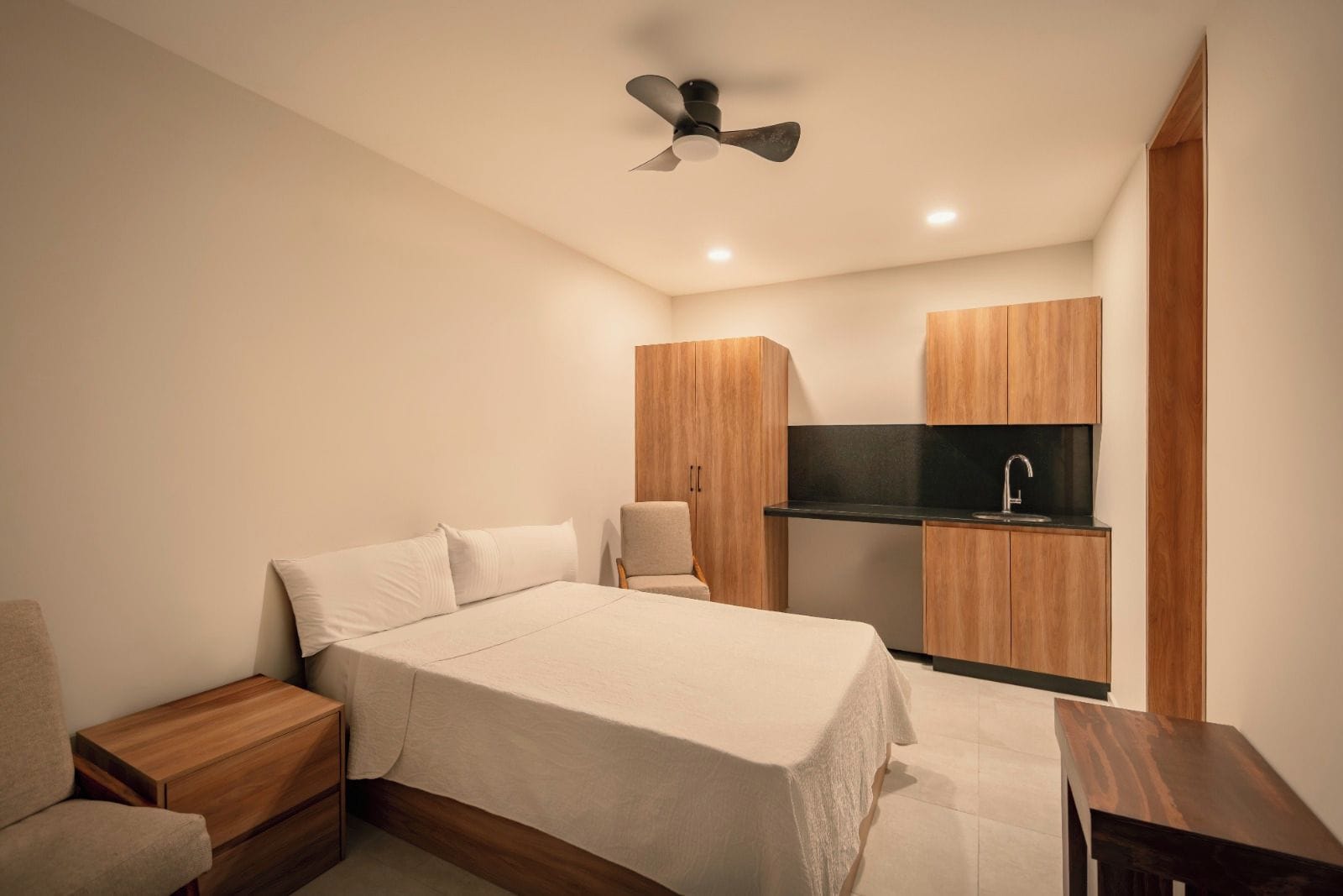 A bedroom with a bed, bedside tables, a chair, a ceiling fan, and a kitchenette area with a sink and cabinets.