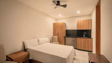 A bedroom with a bed, bedside tables, a chair, a ceiling fan, and a kitchenette area with a sink and cabinets.