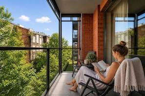 Ein Balkon mit Blick auf einen Garten und Gebäude, eine Person liest ein Buch, und ein Blumentopf.