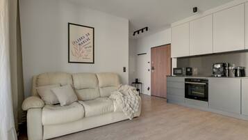 Ein modernes Wohnzimmer mit einem beigen Sofa, einem gerahmten Kunstwerk an der Wand und einer Küchenzeile mit Geräten.