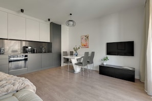Eine moderne Küche mit integrierten Geräten, ein Essbereich mit einem weißen Tisch und grauen Stühlen sowie ein an der Wand befestigter Flachbildfernseher.