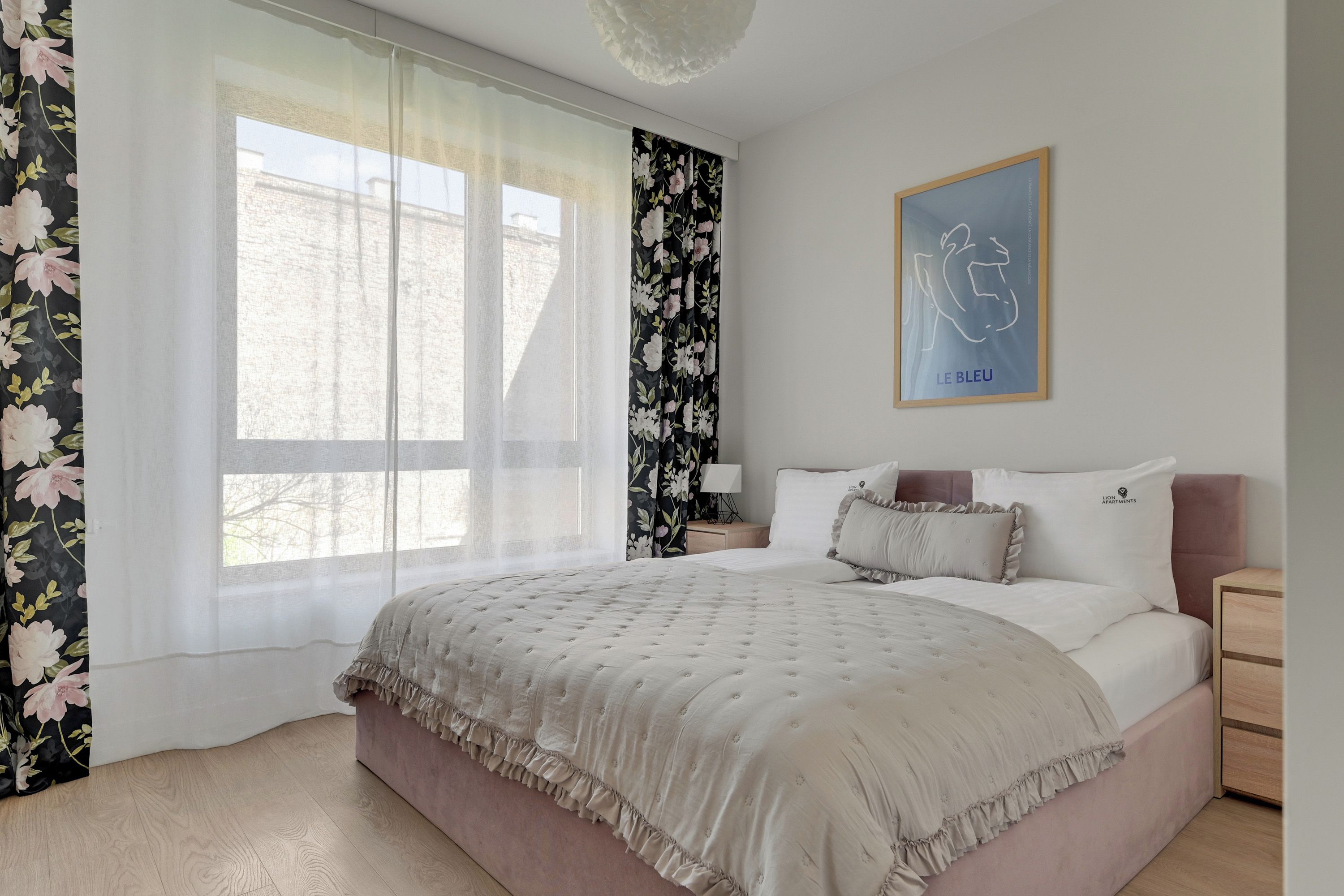 A bedroom with a large window, a bed with a ruffled bedspread, a nightstand with a lamp, and a framed picture on the wall.