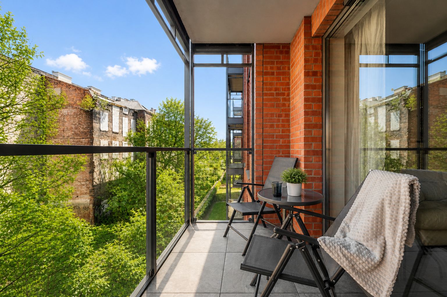 Een balkon met uitzicht op een tuin en gebouwen, voorzien van een tafel, stoelen en een bloempot.