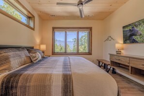 A bedroom with a large bed, a ceiling fan, a window with a mountain view, a bedside table with a lamp, and a wall-mounted TV. - Mountain Views, Hot Tub & Game Room w/ Skee-Ball (Gatlinburg)