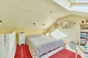 Ein Schlafzimmer mit schrägen Wänden, einem Bett mit floralem Bettzeug, einem Nachttisch, einer Lampe, einem Spiegel und einem Bücherregal.