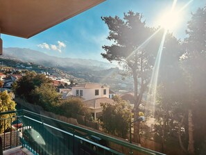 Ein Balkon mit Blick auf eine Hügelstadt, einem klaren Himmel und der Sonne, die durch die Bäume scheint.