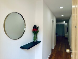 Un couloir avec un miroir rond, une étagère sombre ornée d’un vase de fleurs et une porte dotée d’une poignée ronde.