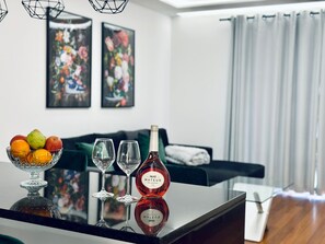 Ein modernes Wohnzimmer mit einem Glastisch, auf dem eine Flasche Mateus Wein, zwei Weingläser und eine Obstschale stehen.