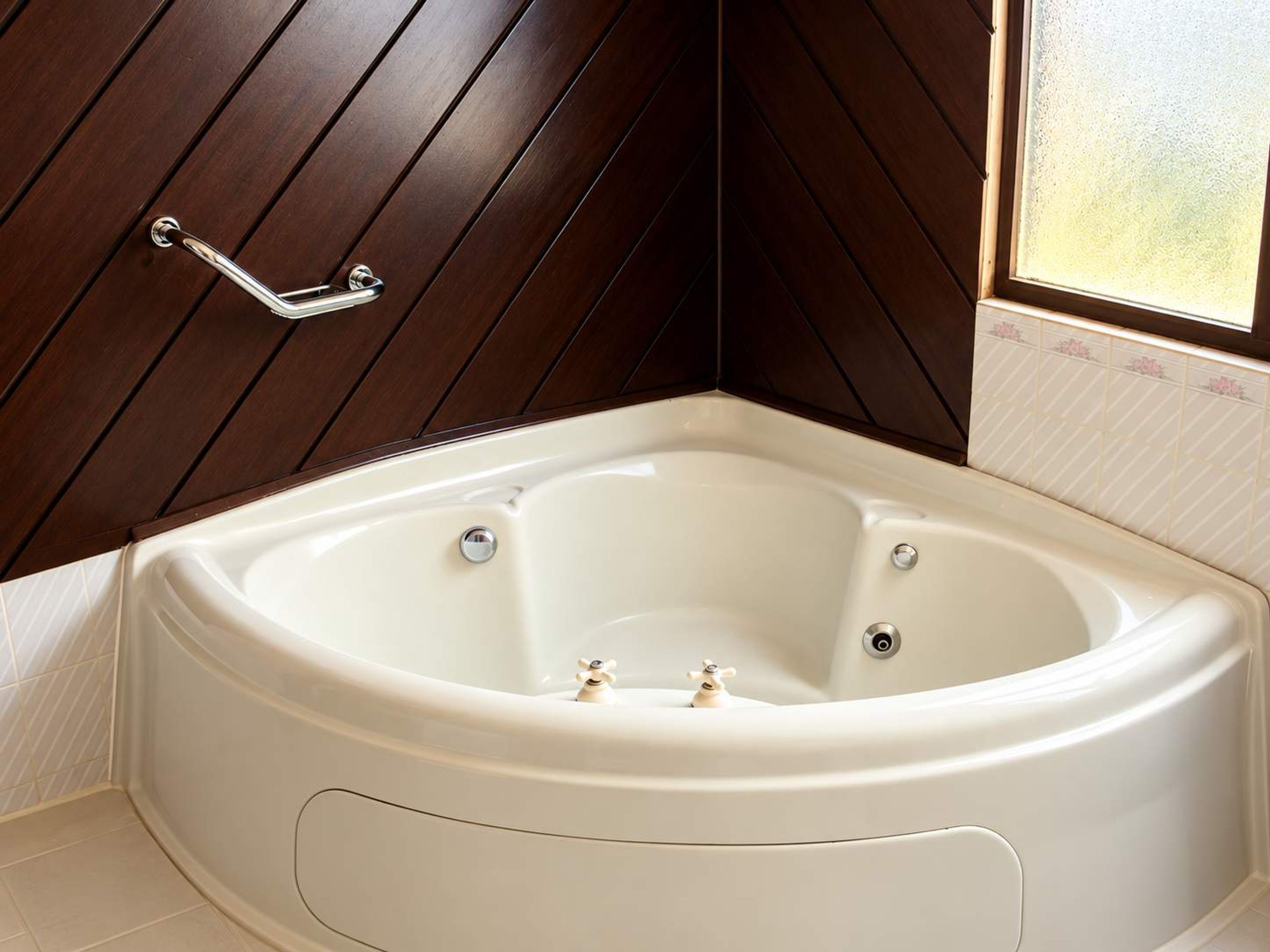 A corner bathroom with a freestanding bathtub, wooden paneling on the walls, and a window with frosted glass.