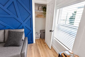 A room with a blue accent wall, a grey sofa, a wooden floor, a white door with blinds, and a laundry basket. - Historic Loft - 2 I NRG   MedCenter   DTown   FIFA (Houston)
