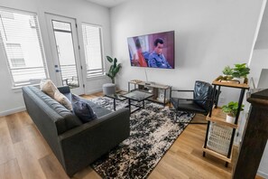 Uma sala de estar moderna com um sofá, mesa de centro e televisão. Há janelas com persianas, uma planta e uma pequena mesa com objetos.