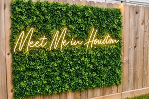 Ein Holzzaun mit einem üppigen Grünstreifen und einem Neon-Schild mit der Aufschrift 'Meet Me in Houston'.