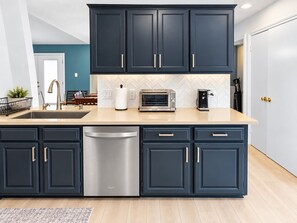 A modern kitchen with dark cabinetry, a stainless steel dishwasher, a built-in oven, and a coffee maker. - 3BR Home with Firepit I Easy Access to NRG   FIFA (Houston)
