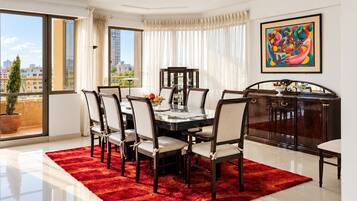 A dining area with a round table, six chairs, a red rug, a city view through large windows, and a colorful abstract painting on the wall.