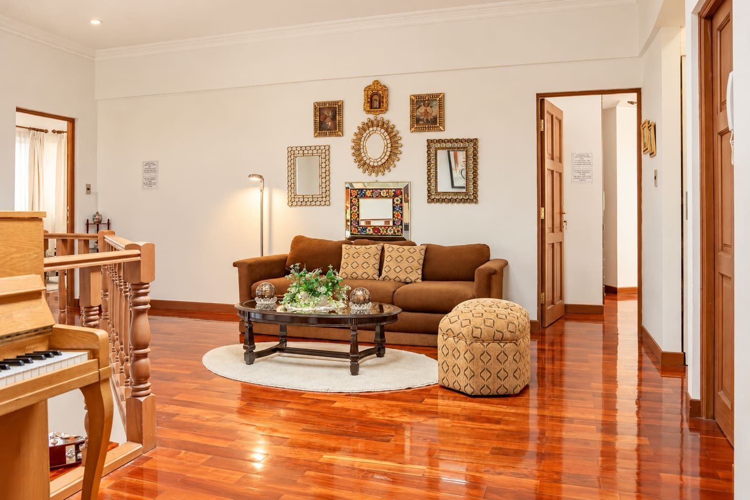 A living room with a brown sofa, a patterned ottoman, a coffee table with decorative items, a piano, and a wall with framed artwork.