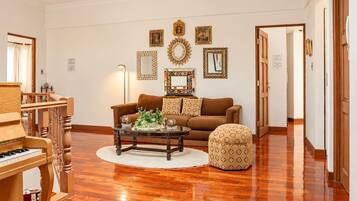A living room with a brown sofa, a patterned ottoman, a coffee table with decorative items, a piano, and a wall with framed artwork.